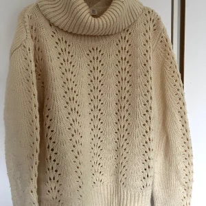 Beige stickad polotröja med hålmönster - Supermysig beige stickad polotröja med bred krage och snyggt hålmönster framtill. Tröjan har lång ärm och ribbade muddar. Perfekt för lager-på-lager och chill höst- och vinterstil.