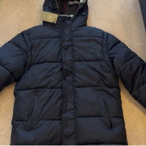  pufferjacka/väst från Burberry - Snygg pufferjacka från Burberry med avtagbara ärmar så du kan bära den som väst. Jackan har huva med rutigt foder, dragkedja och knappar framtill samt två fickor med dragkedja. Perfekt för kalla dagar och riktigt stilren look.
