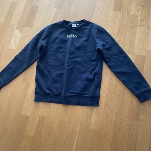 Mörkblå sweatshirt från BOSS - Snygg mörkblå sweatshirt från BOSS med vit logga broderad på bröstet. Tröjan har rund halsringning och långa ärmar, perfekt för en clean och stilren look. Tillverkad i mjuk bomull för maximal komfort.
