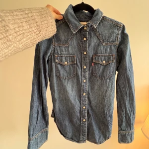 Levi's jeansskjorta med tryckknappar - Snygg jeansskjorta från Levi's i klassisk blå denim med kontrastsömmar och två bröstfickor med tryckknappar. Skjortan har lång ärm, spetsig krage och western-inspirerade detaljer. Säljs inte i butik eller på nätet längre. 