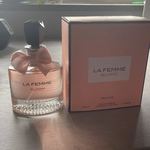 La Femme Bloom parfym 100ml - La Femme Bloom från RiiFFS är en elegant parfym i rund glasflaska med ljusrosa doft och söt rosett vid halsen. Kommer i en matchande ljusrosa kartong. Flaskan rymmer 100ml och har en svart, multifasetterad kork. Perfekt för dig som gillar feminina och fräscha dofter. Cirka 100 ml kvar 