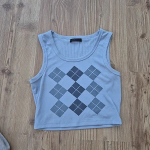 Ljusblå ribbad linnetopp med rutor - Supersnygg ljusblå croppad linnetopp med ribbad struktur och rutigt argylemönster i mörkblått och grått framtill. Ärmlös modell med rund halsringning, perfekt för en trendig och sportig look.