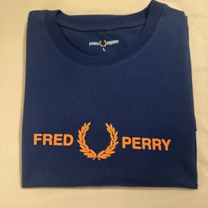 Blå Fred Perry t-shirt med tryck - Helt ny och oanvänd marinblå t-shirt från Fred Perry med orange logga och text framtill. Klassisk rund hals och korta ärmar. Tillverkad i mjuk bomull som känns skön mot huden. Perfekt för dig som gillar stilrena och sportiga plagg. Liten i storlek och passar M.