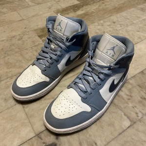 Nike Air Jordan 1 Mid Blå/Vit - Nike Air Jordan 1 Mid i blått och vitt skinn med klassisk snörning och perforerad tå. Snygg retrostil med högt skaft och ikonisk swoosh på sidan. Perfekta för dig som vill sticka ut med en tidlös sneaker från Jordan-serien. Har du någon fråga så kontakta gärna🙂