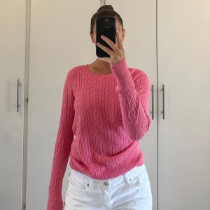 Rosa kabelstickad tröja - Säljer en rosa kabelstickad tröja med rund halsringning och långa ärmar. Tröjan har en klassisk kabelstickad struktur och är i en snygg, ljusrosa färg. Perfekt att styla med jeans eller kjol för en fräsch look. Knappt använd 