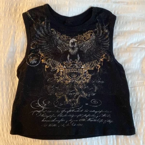 Svart croppad linne med örntryck - Cool svart croppad linne med stort örntryck och texten 'Bike Club' framtill. Trycket har detaljer i guld och vitt, med snygga mönster och skrivstil längst ner. Ärmlös modell med rund halsringning, perfekt för en edgy look 🫶🫶