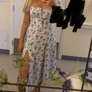 Blommig vit och blå långklänning - Supersöt långklänning med blått blommönster på vit botten. Klänningen har snörning framtill man kan ställa in själv, dragkedja i sidan, slits och en smickrande v-ringning. Perfekt för sommarens alla tillfällen och ger en romantisk vibe.