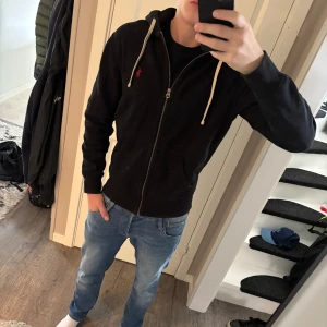 Svart hoodie från Polo Ralph Lauren - Svart hoodie från Polo Ralph Lauren med dragkedja, vit snörning i huvan och röd broderad logga på bröstet. Klassisk passform och mjukt material, perfekt för chill dagar. Snygg och enkel att matcha med jeans eller joggers. Storlek s helt ny