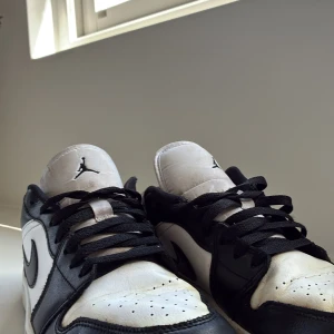 Nike Air Jordan 1 Low svart/vit - Snygga Nike Air Jordan 1 Low sneakers i svart och vitt skinn med klassisk Jordan-logga på tungan och broderad Air Jordan-vinge på hälen. Lågt skaft, svarta snören och perforerad tå för extra stilpoäng. Perfekta för dig som gillar streetwear och ikoniska sneakers.