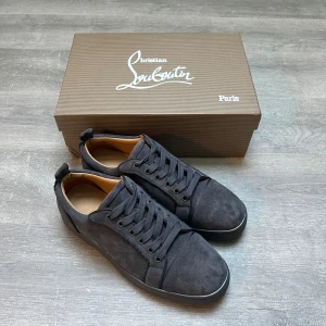 Christian Louboutin svarta sneakers mocka - Snygga svarta sneakers från Christian Louboutin i mjuk mocka med klassisk röd sula och diskret logga på insidan. Skorna har rund tå, platt sula och svarta snören. Perfekta för dig som vill ha en lyxig och stilren look med en ikonisk touch.