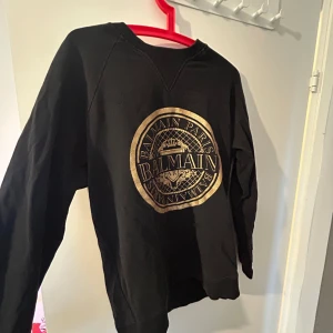 Balmain sweatshirt - Svart sweatshirt från Balmain Paris med stort, glansigt guldfärgat logotryck på bröstet. Tröjan har rund halsringning och långa ärmar. Perfekt för dig som gillar exklusiva streetwear-vibbar och vill sticka ut med en ikonisk look.