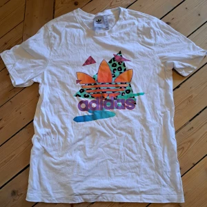 Vit Adidas t-shirt med färgglatt tryck - Vit t-shirt från Adidas med stort färgglatt tryck på bröstet. Trycket har leopardmönster, orange och turkosa detaljer samt lila Adidas-logga. Klassisk rund hals och korta ärmar. Skön bomullskvalitet, perfekt för dig som gillar streetstyle, den är en använd 1-2 gånger.