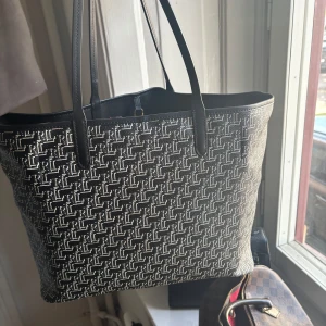 Svart shoppingväska från Lauren Ralph Lauren - Snygg svart shoppingväska från Lauren Ralph Lauren med vitt RL-mönster över hela väskan. Väskan har långa svarta handtag, guldfärgade metalldetaljer och dragkedja på insidan. Tillverkad i tålig syntet med läderdetaljer. Perfekt för dig som vill ha plats med allt.
