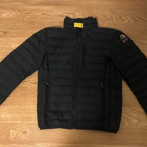 Parajumpers dunjacka - Säljer en svart dunjacka från Parajumpers med klassisk patch på ärmen och gul detalj vid kragen. Jackan är superlätt, har tre dragkedjeförsedda fickor och quiltad design. Insidan är grå och jackan har hög krage för extra skydd mot kylan.