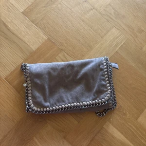 Stella McCartney liknande väska  - Säljer en snygg beige axelväska i mocka med silverfärgad kedja som axelrem och runt kanterna. Väskan har magnetlås, innerfack med dragkedja och en rund metallbricka som detalj. Perfekt storlek för det viktigaste och ger en cool touch till outfiten.