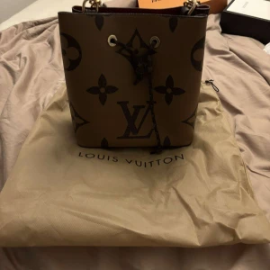 Louis Vuitton Monogram Bucket Bag - Säljer en ikonisk bucket bag från Louis Vuitton med klassiskt monogrammönster i brunt och beige. Väskan har guldfärgade metalldetaljer, snörning upptill och röd insida. Tillverkad i exklusivt skinn och har både axelrem och handtag. Perfekt för dig som vill sticka ut med lyxig stil.
