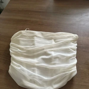 Vit bandeau tubtopp med korsett - Säljer en snygg vit bandeau tubtopp med draperad front och snörning i ryggen. Det är en typ av korsett. Toppen har en cool omlottkänsla och är gjord i ett tunt, lätt material som känns lyxigt. Perfekt för dig som vill ha en trendig och stilren look. 💕