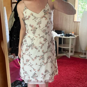 Vit blommig sommarklänning med smala axelband - Superfin vit kortklänning med smala axelband och v-ringning. Klänningen har ett romantiskt blommönster i rosa och beige toner, markerad midja och lätt, luftig känsla. Perfekt för varma dagar och ger en fräsch vibe.