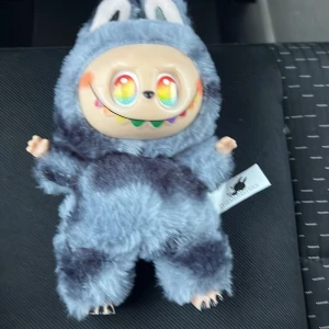 Secret labubu  - Supersöt och unik ryggsäck i form av ett monster med regnbågsfärgade ögon och tänder. Väskan är täckt av mjuk, blå och mörkblå pälsimitation och har ett praktiskt fäste upptill. Perfekt för dig som vill sticka ut med en lekfull accessoar.