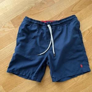 Marinblå badshorts Polo Ralph Lauren för barn ålder 14-16 - Snygga marinblå badshorts från Polo Ralph Lauren med vit snörning i midjan och röd broderad logga på benet. Shortsen har elastisk midja, innerbyxa i mesh och en bakficka med dräneringshål. Perfekta för stranden eller poolen!