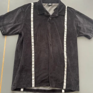 Svart pikétröja i sammet Sweet SkTbs - Svart pikétröja från Sweet SkTbs i mjuk sammet. Tröjan har korta ärmar, klassisk krage med knapp och vita band med logga längs fram- och baksidan. Perfekt för dig som gillar unika detaljer och skön känsla. Storlek S men har en baggy look