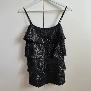 Svart paljettlinne med volanger - Glittrig svart linnetopp med tunna axelband och flera lager volanger. Hela toppen är täckt av svarta paljetter som ger en riktigt festlig look. Perfekt för dig som vill sticka ut och glänsa på nästa event.
