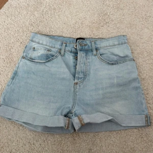 Ljusblå jeansshorts från 157 - Ljusblå jeansshorts från 157 med klassisk femficksdesign, uppvikta benslut och hög midja. Shortsen har bälteshällor, knapp- och dragkedjestängning samt snygga bakfickor. Perfekta för varma dagar och enkel att matcha med allt.
