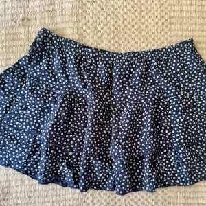 Blommig blå kjol från Brandy Melville - Supersöt kort kjol från Brandy Melville i marinblått med små vita blommor. Kjolen har volangdetaljer och resår i midjan för en bekväm passform. Perfekt för dig som gillar en feminin och trendig stil.