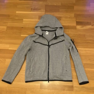Grå Nike Tech  - Snygg grå vindjacka från Nike med svart dragkedja och detaljer. Jackan har huva, långa ärmar och två fickor framtill. Perfekt för dig som gillar sportig stil och vill ha något lätt och smidigt att slänga på dig.