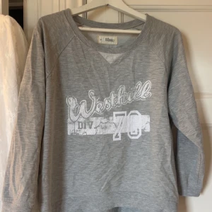 Grå sweatshirt Westhill DIV 70 - Grå sweatshirt från Rebel med tryck 'Westhill DIV 70' i vitt framtill. Tröjan har v-ringning, långa ärmar och är tillverkad i mjuk bomullsmix. 