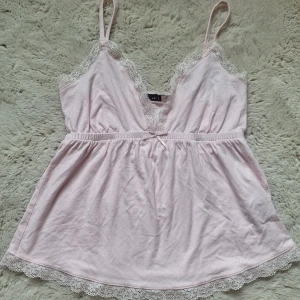 Ljusrosa spetslinne från Nelly XS - Supersött ljusrosa linne från Nelly med smala axelband och v-ringning. Toppen har fina spetsdetaljer längs urringningen och nederkanten samt en liten rosett framtill. Perfekt för en romantisk look eller som layering-piece.