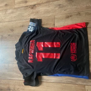 FC Barcelona Raphinha #11 Nike tröja - Snygg svart FC Barcelona fotbollströja med Raphinha och nummer 11 på ryggen. Tröjan har röda och blå detaljer på sidorna, Champions League-märke på ärmen, Spotify-logga på bröstet och klubbmärke i svart. Tillverkad i lätt och ventilerande material. Skickar inom 24 timmar