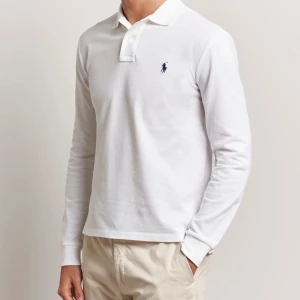 Vit långärmad pikétröja Polo Ralph Lauren - Vit långärmad pikétröja från Polo Ralph Lauren med klassisk krage och knäppning framtill. Tröjan har broderad marinblå logga på bröstet och är gjord i mjuk bomull. Perfekt för en clean och stilren look.