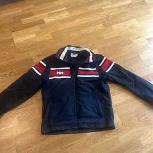 Helly Hansen vindjacka - Snygg marinblå vindjacka från Helly Hansen med röda och vita ränder på bröstet och ärmarna. Jackan har dragkedja framtill, ståkrage och två fickor med dragkedja. Perfekt för dig som gillar sportig och stilren look.