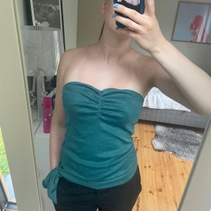 Glittrig grön bandeau tubtopp - Säljer en snygg grön bandeau tubtopp med glittriga detaljer och rynk framtill. Toppen är axelbandslös och har en söt knytning i sidan. Perfekt för dig som vill sticka ut lite extra i sommar!