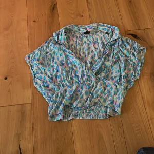 Färgglad omlottblus från Lindex - Säljer en superfin omlottblus från Lindex i storlek M. Blusen har ett färgstarkt mönster i blått, rosa, grönt och vitt, med korta ärmar och resår i midjan för en snygg passform. Perfekt för dig som gillar färg och vill sticka ut lite extra!