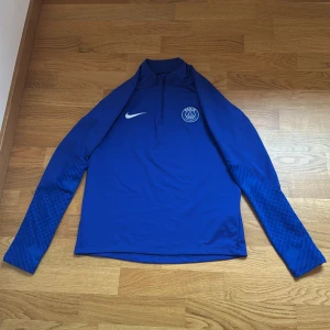Blå långärmad PSG träningströja Nike - Snygg blå långärmad träningströja från Nike med PSG-logga på bröstet och diskret mönster på ärmarna. Slim fit-modell med halv dragkedja framtill. Perfekt för fotbollsträning eller när du vill representera Paris Saint-Germain.