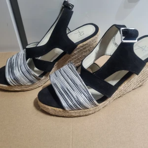 Svarta kilklack sandaler med silverdetaljer - Snygga sandaler med svart ovandel och breda remmar i silver med randigt mönster. Skorna har kilklack i flätad jute och justerbart spänne vid ankeln. Perfekta för sommaren och ger en cool touch till din outfit.