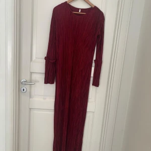 Plisserad vinröd långklänning - Vinröd långklänning med plisserat tyg och långa ärmar. Klänningen har en rak passform och rund halsringning. Ärmarna har en liten detalj med sydd kant vid armbågen. Tyget har en glansig finish som ger en lyxig känsla.