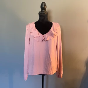 Rosa blus med volang från Sisters Point - Supersöt ljusrosa blus från Sisters Point med långa ärmar och v-ringning. Blusen har en snygg volangdetalj runt halsen som ger en romantisk vibe. Passar perfekt inför i sommar!