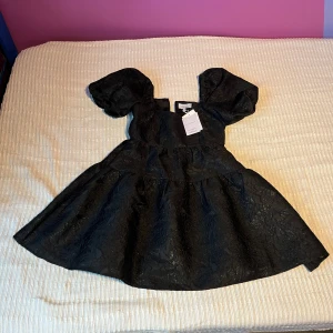 Svart puffärmad klänning & Other Stories - Supersnygg svart kortklänning från & Other Stories Paris Atelier med puffärmar och markerad midja. Klänningen har ett subtilt blommönster i tyget och en lätt glansig finish. Perfekt för dig som vill sticka ut med en cool och elegant look.