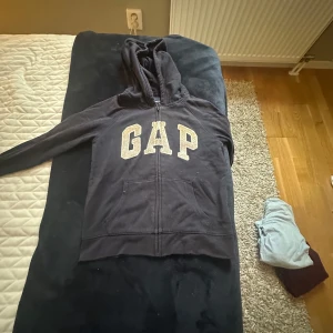 Svart hoodie med paljetter från GAP - Svart hoodie från GAP med dragkedja och huva. Stora bokstäver på bröstet dekorerade med paljetter. Köpt secondhand och är i okej skick, inga defekter riktigt. Storlek 12-13 år💕