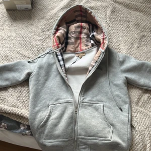 Grå hoodie från Burberry med dragkedja - Säljer en grå hoodie från Burberry med klassiskt rutigt foder i huvan och broderad logga på bröstet. Tröjan har dragkedja, snörning i huvan och två stora fickor framtill. Mjuk insida och ribbade muddar för extra komfort.