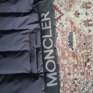 Svart Moncler väst stl S - Säljer en svart pufferjacka från Moncler med tydlig logga på bröstet och bredt Moncler-band längs sidan. Jackan har en stilren, quiltad design och är tillverkad i syntetmaterial. Perfekt för dig som vill ha en clean och exklusiv look.