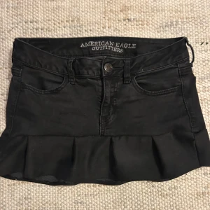Svart jeanskjol med volang från American Eagle - Supersnygg svart omsydd jeanskjol från American Eagle med volangkant nertill. Klassisk femficksmodell, bälteshällor och knappstängning framtill. Kjolen är kort och har en figurnära passform som ger en cool och trendig look.