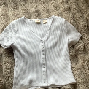 Vit ribbad topp med knappar - Superfin vit ribbad topp från Levi's med korta ärmar och v-ringning. Toppen har knappar hela vägen framtill och är i ett mjukt, stretchigt material som sitter snyggt på kroppen. Perfekt till jeans eller kjol för en clean look.