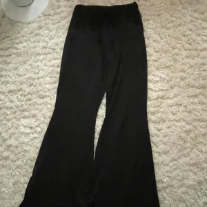 Svarta vida byxor i satinlook - Säljer ett par svarta byxor med snyggt fall och breda ben. De har en hög midja och är gjorda i ett glansigt material som ger en lyxig satinlook. Perfekta om du vill sticka ut lite extra med en trendig och bekväm stil. Har ett litet hål på knäet men det syns knappt