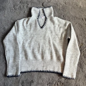 Grå stickad tröja från Gina Tricot - Mysig grå stickad tröja från Gina Tricot med mörkblå virkade detaljer längs v-ringningen och nederkanten. Tröjan har lång ärm och ribbad mudd nedtill, perfekt för lager på lager under kyliga dagar.