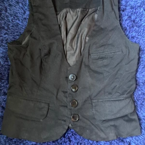 Svart randig väst från Vero Moda - Klassisk svart väst från Vero Moda i storlek 38 med diskreta tunna ränder. Västen har fyra knappar framtill, två ”fickor” (ej funktionella). 