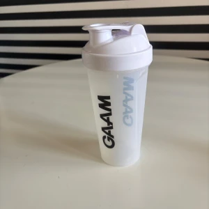 Vit GAAM shaker 800 ml - Säljer en vit shaker från GAAM med svart logga, rymmer 800 ml. Perfekt för träning och smoothies, med praktiskt lock och sil för att blanda drycker smidigt. Enkel att ta med till gymmet eller skolan.